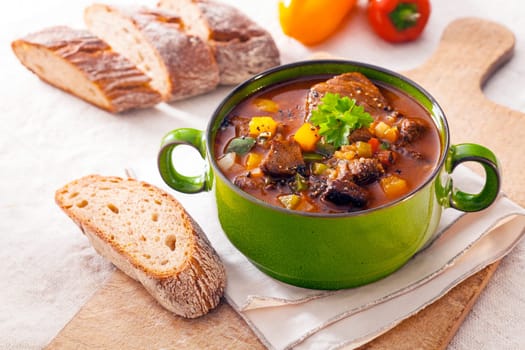 Tasty winter stew in a green metal pot with meat and assorted vegetables in a rich gravy served with fresh crusty bread on a wooden chopping board