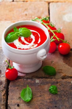 Small bowl of colourful red tomato soup with cream and basil on old weathered bricks with scattered fresh ingredients