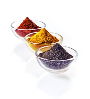 Three clear glass bowls of colourful of ground spice and black lava salt in conical heaps arranged in a receding row on a white background