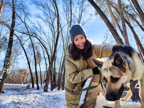 A girl walks cheerfully with her German Shepherd dog through a snowy park surrounded by tall trees on a bright winter day.