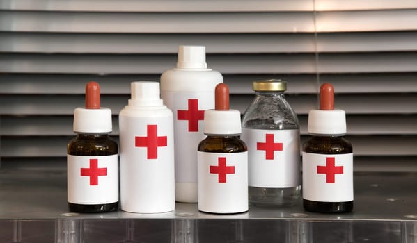 Collection of pharmaceuticals with blank white labels showing a red cross including glass dropper bottles, plastic containers and a glass bottle for liquid for injection