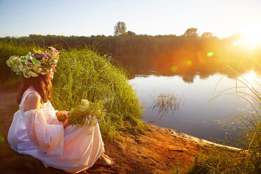 Slavic plump chubby girl in long white dress on the feast of Ivan Kupala with flowers and water in a river or lake on summer evening