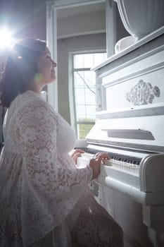 A woman wearing a delicate dress gracefully plays a white piano, illuminated by warm sunlight streaming through a large window.