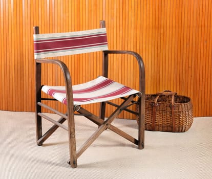 Furniture Still Life of Folding Retro Beach Chair with Striped Pattern in Room with Woven Basket, Beige Carpet and Wood Wall Paneling