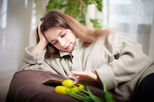 A woman comfortably lies on a couch, engaged with her smartphone while beautiful flowers add a touch of nature to her relaxing space.