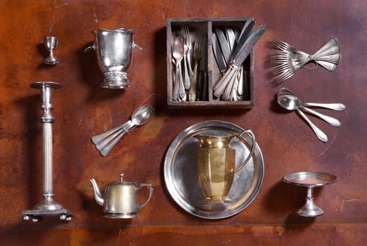 Assorted silver kitchenware arranged on wood with cutlery, candlestick, ice bucket, egg cup, teapot tray and pitcher or jug viewed from above