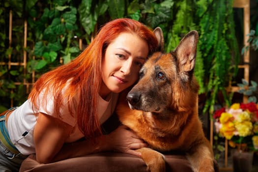 Joyful woman and her large German Shepherd share a loving moment inside a well-decorated room filled with plants.