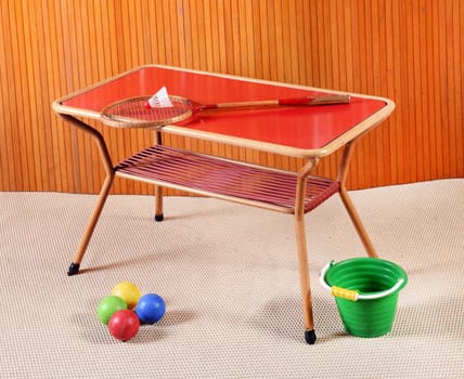 Still Life of Small Vintage Games Table with Badminton Racket, Multi Colored Balls and Green Bucket in Room with Wood Wall Paneling and Beige Carpet