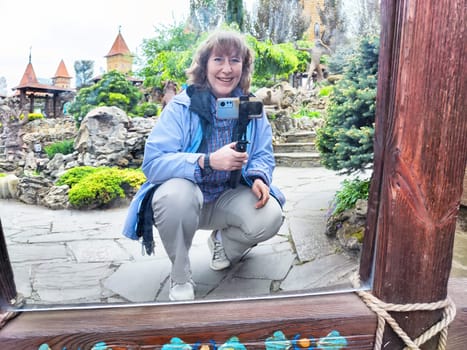 Middle-aged woman takes a humorous selfie in a warped mirror surrounded by lush greenery in a park setting.