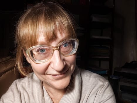 A 50-year-old woman with blonde hair smiles playfully while taking a selfie, enjoying her relaxed moment at home.