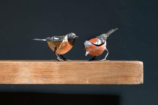 Two vintage metal toy birds on a wooden shelf, one perched and one pecking at the ground in a close up side view against black