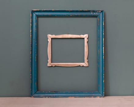 Two empty vintage picture frames, one painted blue, the other a smaller carved frame hanging on a teal colored wall for interior decor