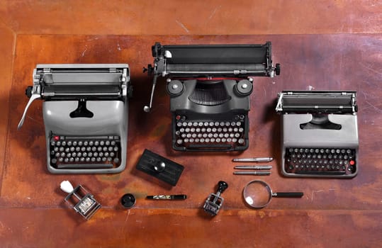 High Angle Still Life View of Three Typewriters with Magnifying Glass, Rubber Stamps, Pens and Other Stationary Objects on Wood Table Surface
