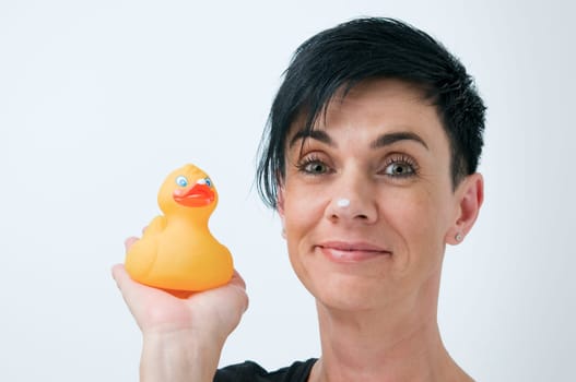 Head-and-shoulder view of a black-haired middle-aged woman against a white background with a yellow rubber duck held next to her,looking friendly into the camera,each with a cream swab on her noses