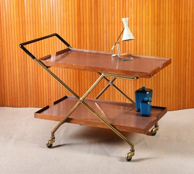 Still Life of Retro Service Trolley on Wheels with Desk Lamp and Blue Jar Set in Room with Beige Carpet and Wood Wall Paneling