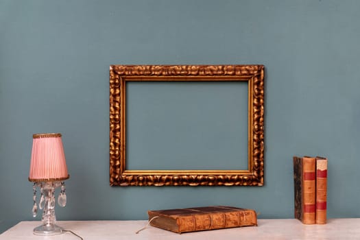 Empty gold rectangular vintage frame hanging on a green wall above a shelf with crystal drop lamp and old leather books in an interior decor concept