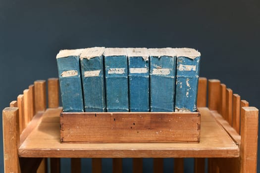 Set of old worn and tattered vintage books with blue spines and labels on a wooden shelf against a grey wall