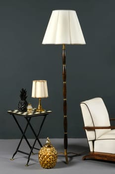 Interior decor with stylish vintage furniture comprising a wooden floor lamp, easy chair and table lamp on a chequered table with cream coloured shades and upholstery against a grey wall