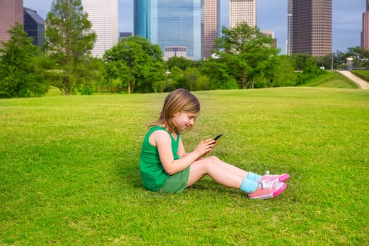 Blond kid girl playing with smartphone sitting on park lawn in city skyline background