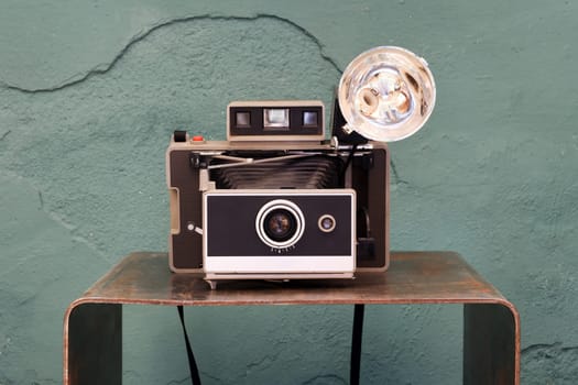 Old vintage instant camera with bellows and lamp on a small shelf in front of a damaged green textured wall in a photographic concept