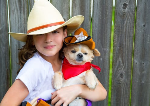 Beautiful cowboy kid girl holding chihuahua dog with sheriff hat in backyard wooden fence