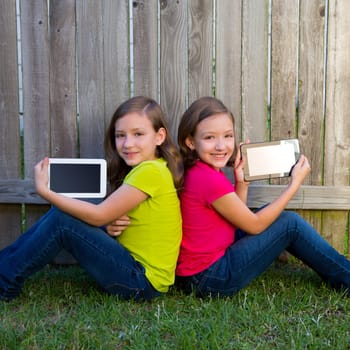Twin sister girls playing with tablet pc sitting on backyard lawn fence leaning on her back