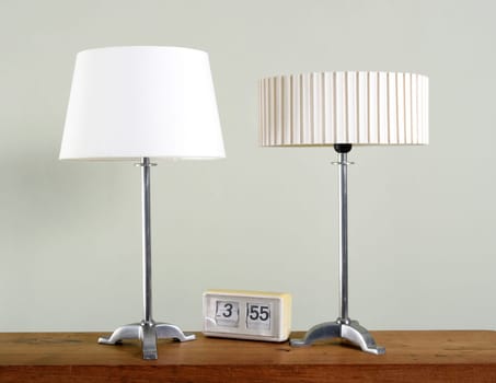 Two vintage metal lamps with different white shades alongside an old clock on a wooden table