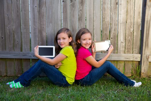 Twin sister girls playing with tablet pc sitting on backyard lawn fence leaning on her back