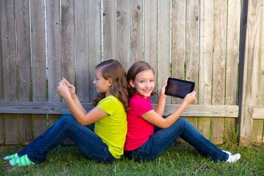 Twin sister girls playing with tablet pc sitting on backyard lawn fence leaning on her back