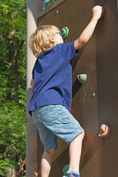 The blond boy climbs the climbing wall in summer day.