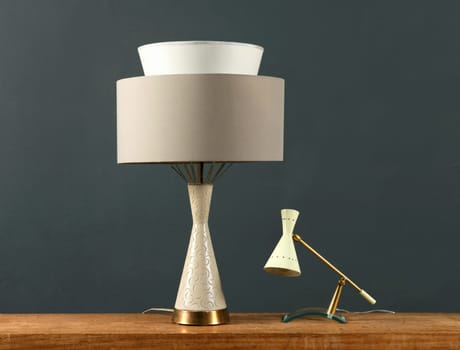 Two vintage lamps on a wood cabinet, one cylindrical with angular base and shade and one small anglepoise lamp against a dark grey wall