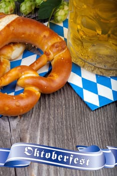 A glass of beer,pretzels and some hop cones on an old rustic wooden table with a blue banner that says Oktoberfest