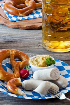 Blue and white paper plate with white sausage,pretzel,mustard and radishes and a glass of beer on an old rustic wooden table