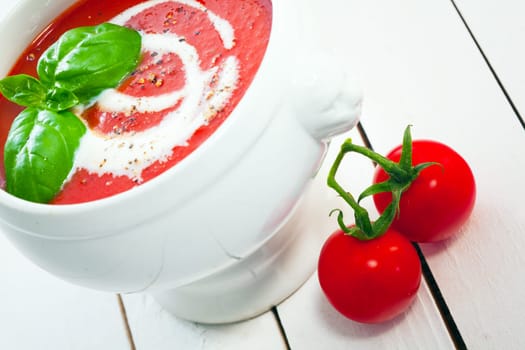 Appetizing bowl of tomato soup drizzled with cream and topped with basil leaves with fresh cherry tomatoes on white painted boards