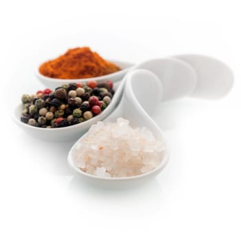 Ceramic spoons filled with natural Himalayan rock salt, whole dried black peppercorns and hot red cayenne pepper on a white background