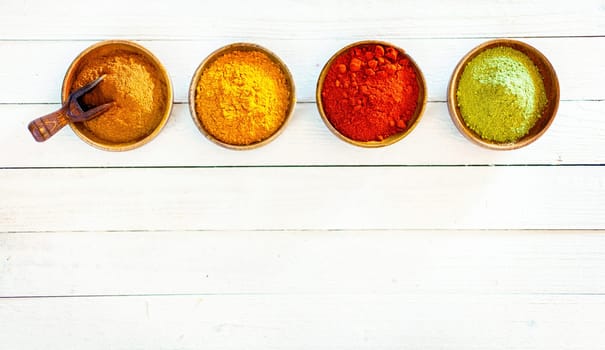 Overhead view of a line of four colourful ground spices in wooden bowls on white painted boards with copyspace below
