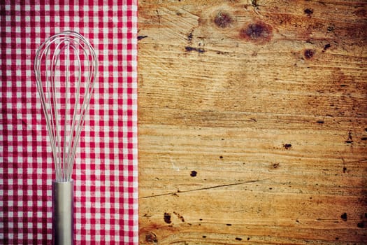Metal cooking whisk, or balloon whisk, lying on a fresh red and white checkered napkin on a wooden countertop in a rustic kitchen with copyspace
