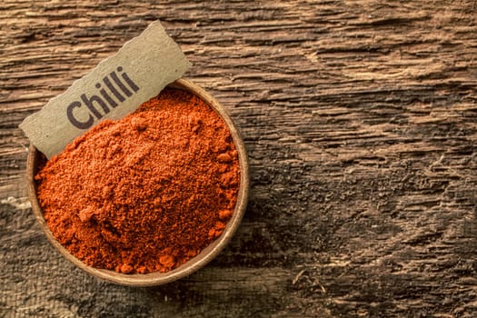 Red hot chilli powder in a rustic wooden bowl with an ornamental name label on a grunge weathered textured wooden surface, overhead view