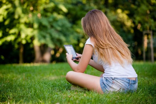 Young woman (teen girl) reading book on electronic book reader - outdoor, rear view