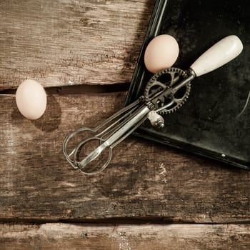 High angle view of two eggs and an old egg beater resting on a baking tray on an old grunge wooden kitchen table
