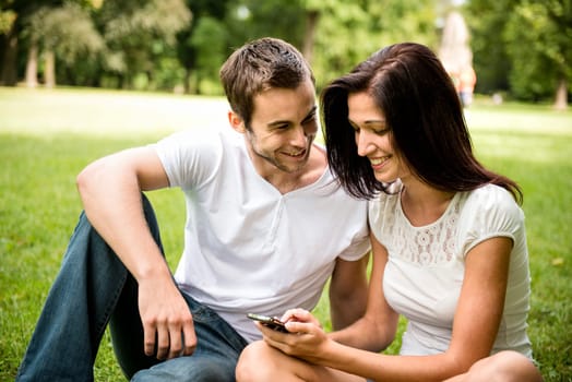 Young happy couple sitting on grass and and looking on smartphone display