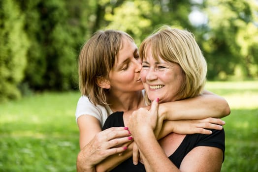Adult daughter kissing her smiling happy senior mother - outdoor in nature