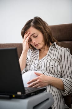 Pregnant woman with headache working on laptop at home on sofa