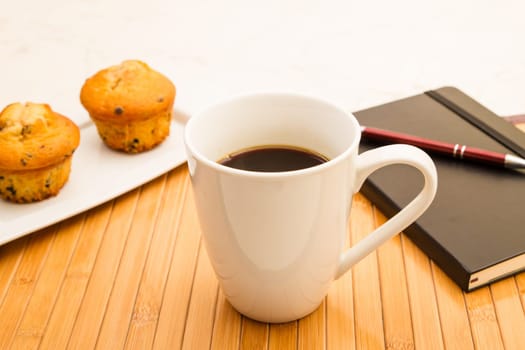 Vanilla with chocolate chips Muffins on a white plate with  a cup of coffee meanwhile taking notes.