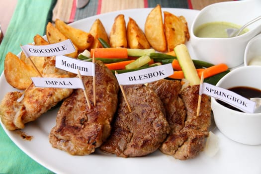 A plate of African game meats with sauces, vegetables, and potatoe fries. Meats are marked with little toothpick signs crocodile, impala, blesbok,springbok