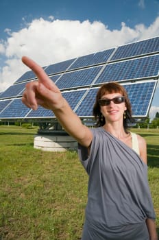Upper body shot of a young,brunette woman with sunglasses in casual casual dress in front of a photovoltaic system on a green meadow in summer points to the top left past the camera