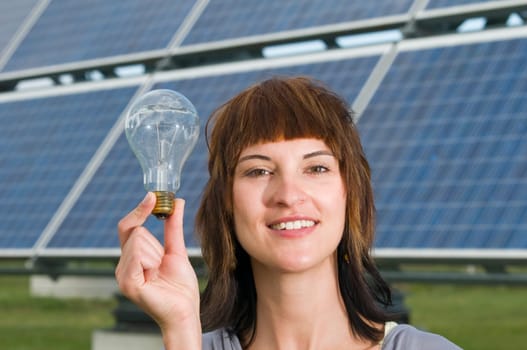 Head-and-shoulder view of a young brunette woman next to her,holding up a conventional light bulb in front of industrial solar panels,smiling into the camera