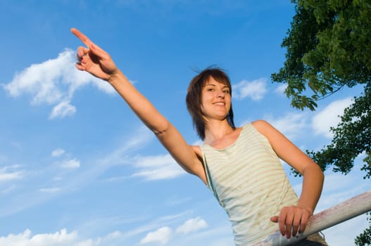 Upper body shot of a young,medium-length brown-haired woman in a sleeveless top standing at a lake in Potsdam (Griebnitzsee) and pointing with her right arm to the side in front of a blue sky with small white clouds