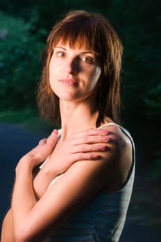 Upper body portrait of a young dark-haired woman in a sleeveless top and crossed arms in front of her chest and hands on her shoulders on a sidewalk in the park at dusk looking into the camera