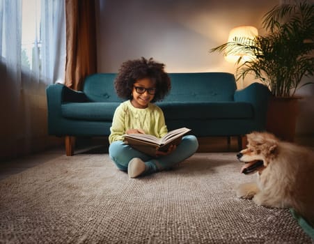 Smiling young Black girl reading book with Shetland Sheepdog in cozy living room.